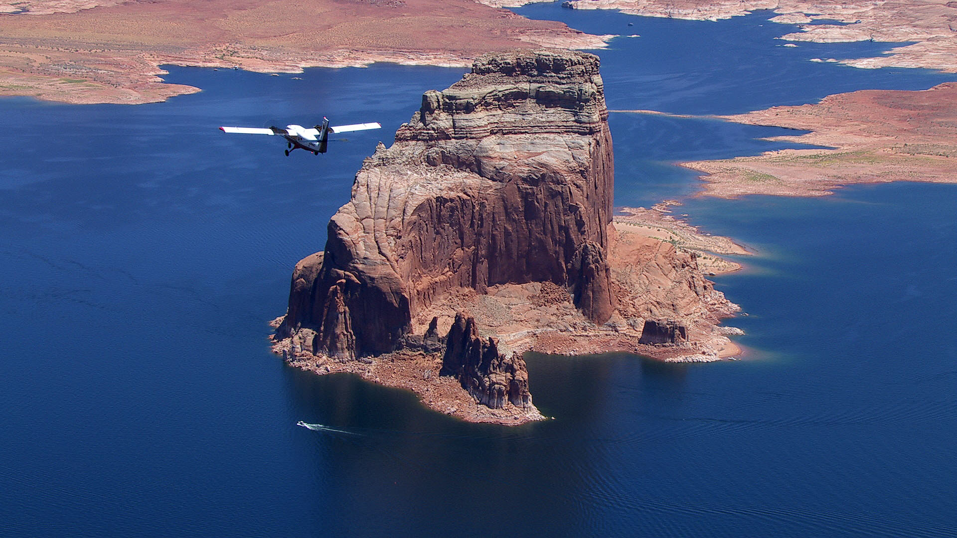 An airplane soars over a blue lake surrounded by desert.