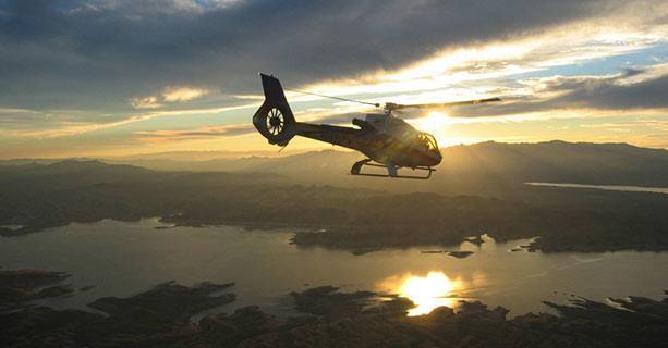 A helicopter flies over Lake Mead as the sun sets in the background.