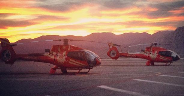 Two helicopters parked on a tarmac with the sun setting behind them.