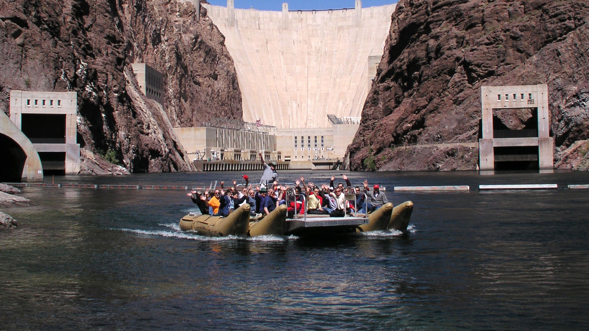 Passengers on a river raft with the Hoover Dam in the background.