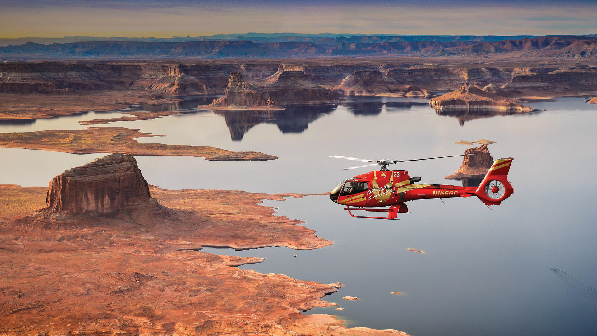 A red helicopter flies over a lake surrounded by desert.