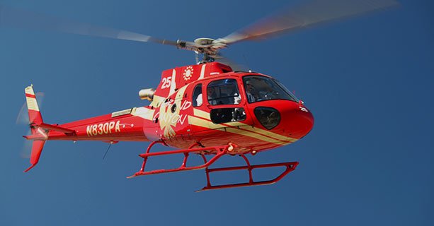 A red EC-130 helicopter hovering in a clear blue desert sky.