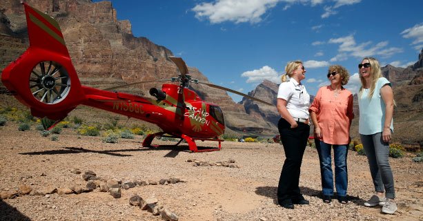 Two female passengers and a female pilot talk in front of a helicopter on the Grand Canyon floor.