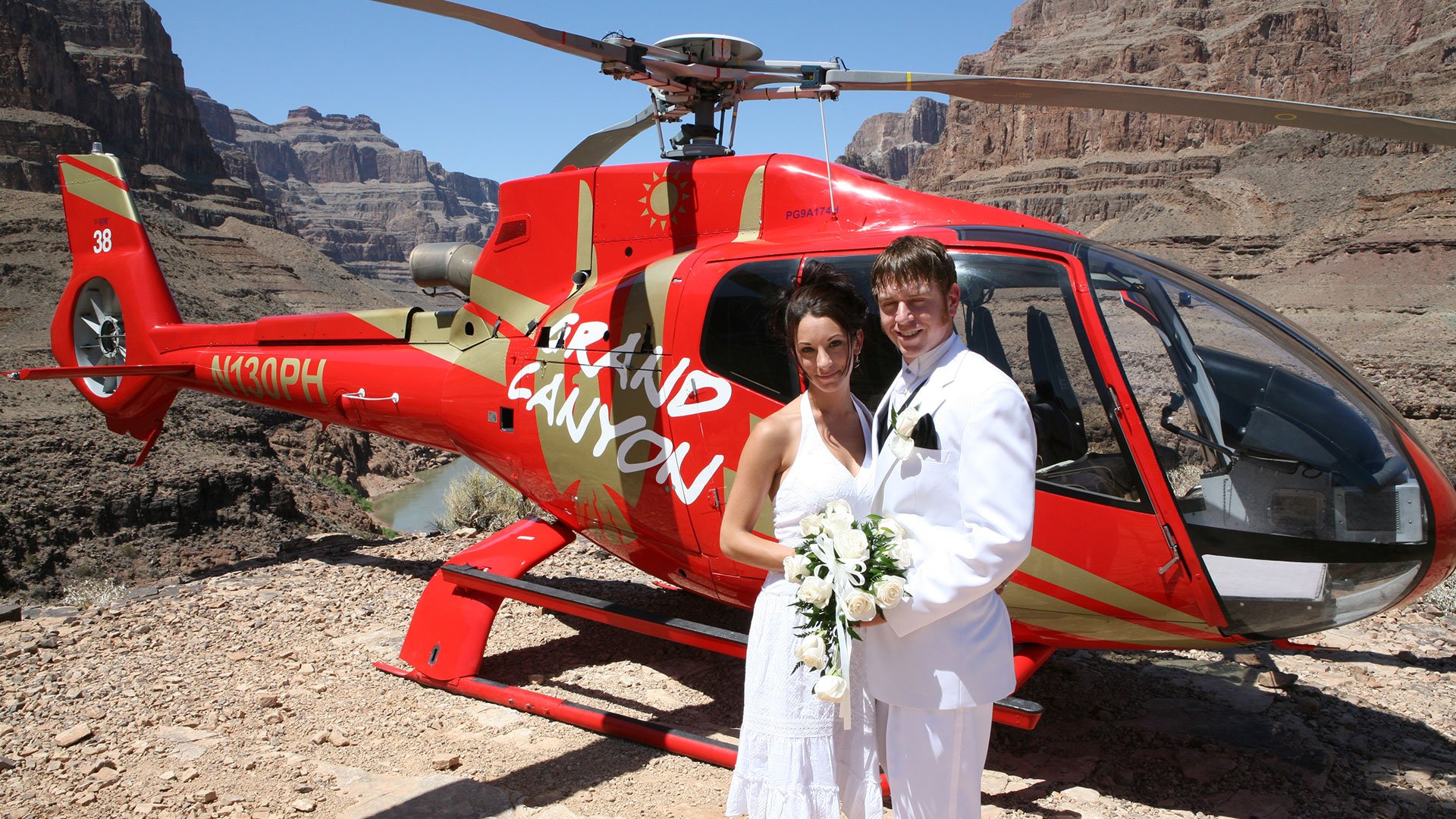 A bride and groom pose in front of a helicopter at the bottom of the Grand Canyon.