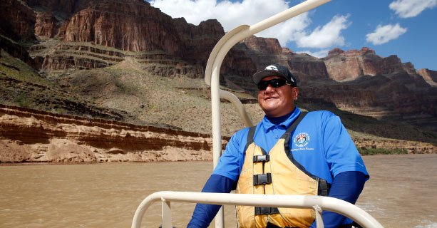 A man steering a pontoon boat along the Colorado River with the Grand Canyon behind him.